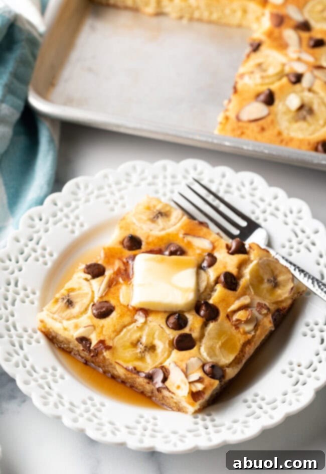 A delicious square of chocolate chip and banana oven baked pancake on a white plate, garnished with butter and syrup, with a fork nearby.