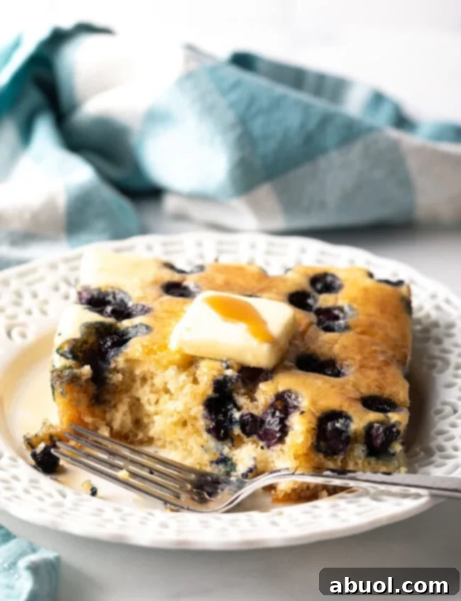 A single square of blueberry baked pancake from the sheet pan, with a bite taken out, served on a white plate with a fork.