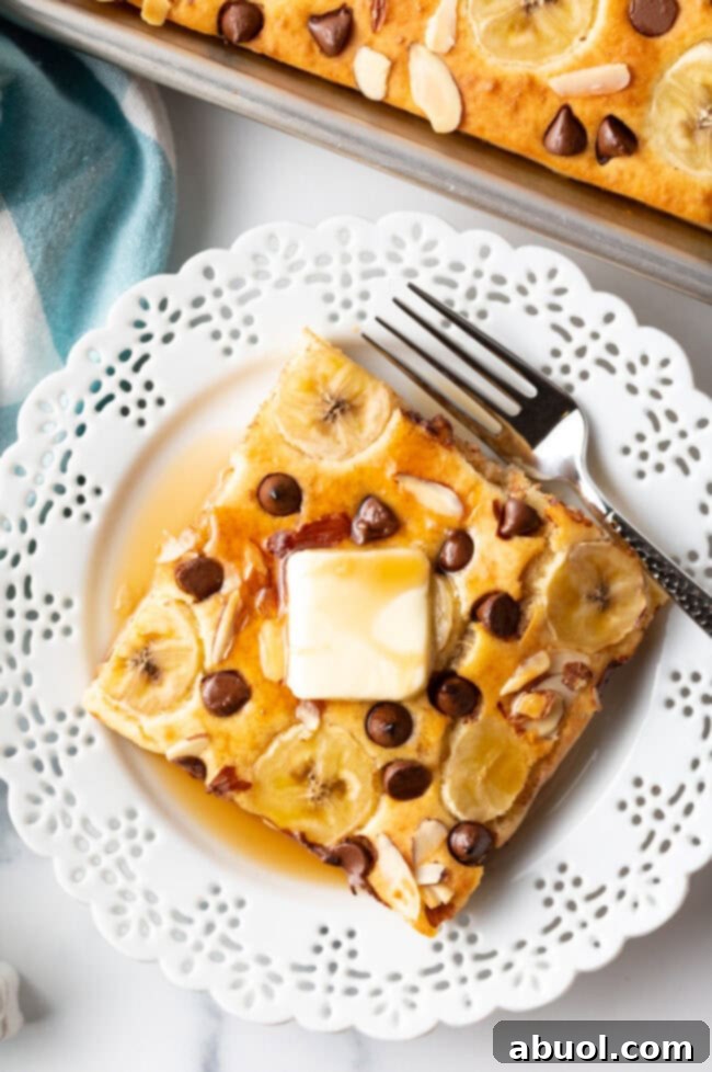 Top-down view of a delicious sheet pan pancake square topped with sliced bananas, chocolate chips, and toasted almond slices, served on a plate with a fork.