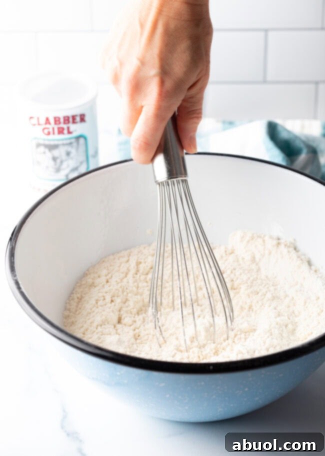 A hand whisking together dry ingredients like flour, sugar, baking powder, and salt in a large blue mixing bowl for sheet pan pancakes.