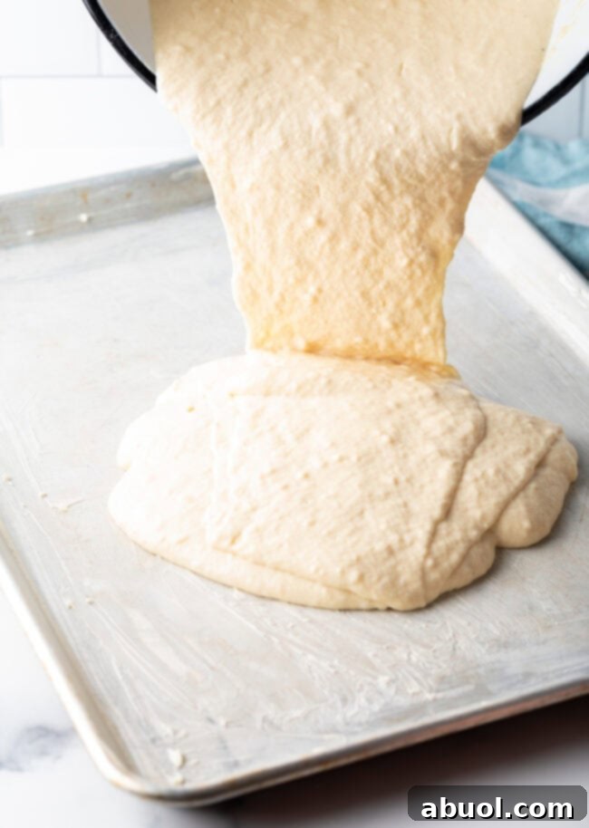 Pouring smooth pancake batter from a mixing bowl into a prepared rimmed baking sheet for oven baked pancakes.