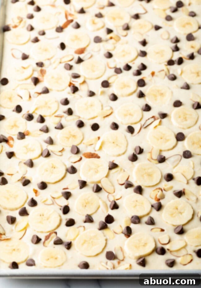 Unbaked pancake batter in a sheet pan, adorned with banana slices, chocolate chips, and almond slivers, ready for the oven.