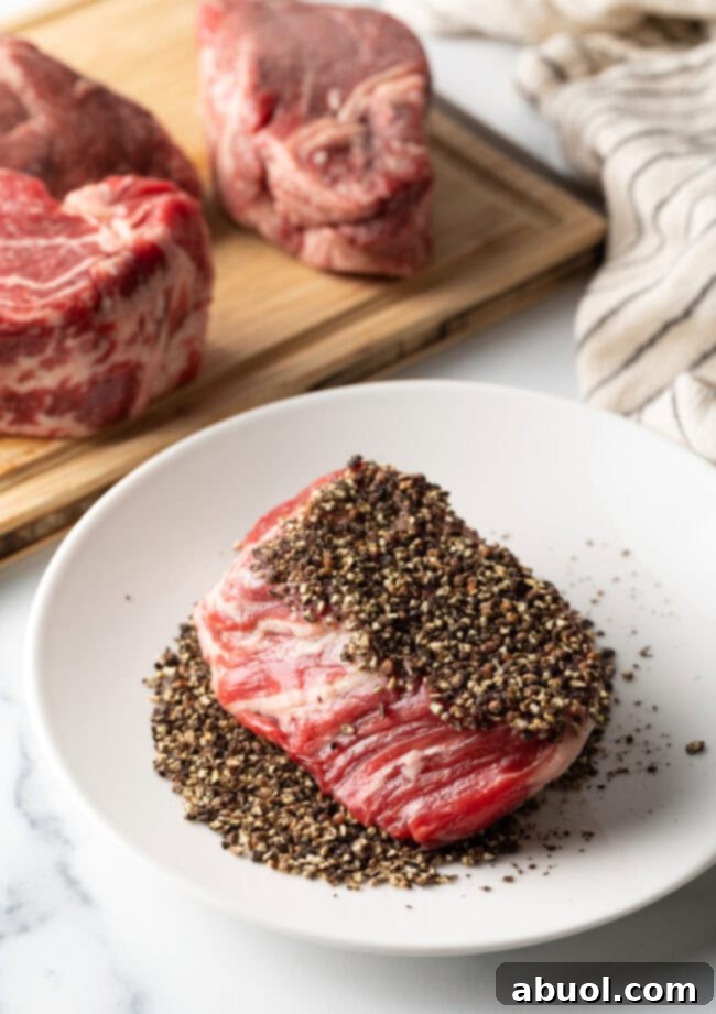 Uncooked beef tenderloin steak being pressed into a plate of coarse black ground peppercorns for the crust.