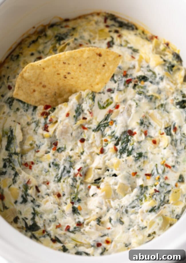 Close-up view of a rich and creamy Crockpot Spinach Artichoke Dip in a slow cooker, with one golden tortilla chip ready for dipping.