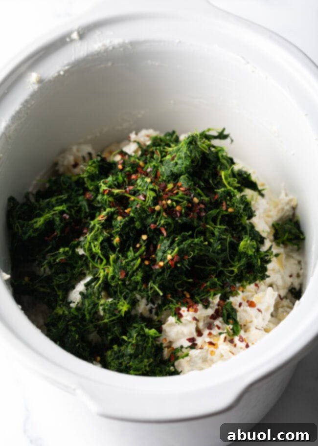 Cooked spinach and seasonings being added to the slow cooker with other dip ingredients.