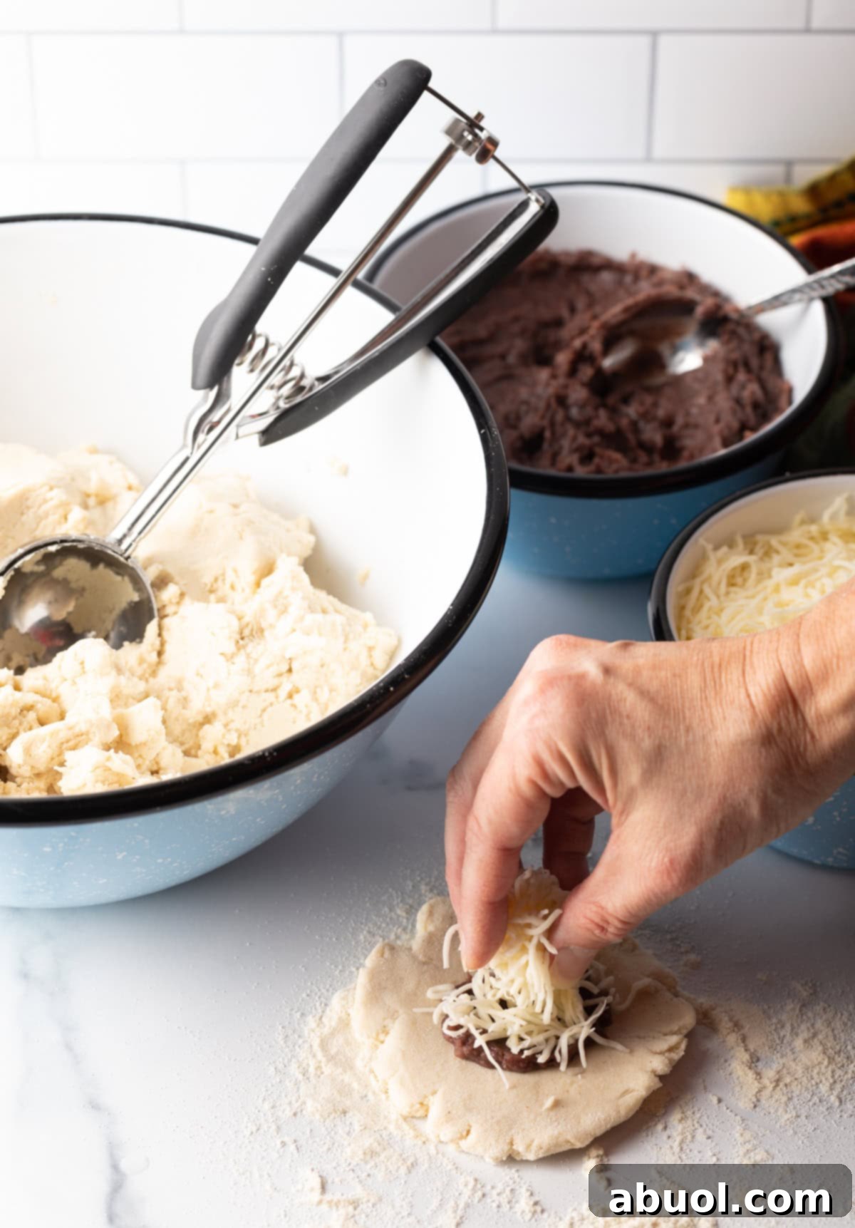 A hand adding a generous amount of shredded Monterey Jack cheese on top of the beans on the flat pupusa dough.