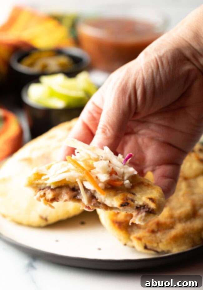 A hand holding a pupusa with a bite taken out, showcasing the melted cheese and beans, beautifully topped with fresh curtido slaw.