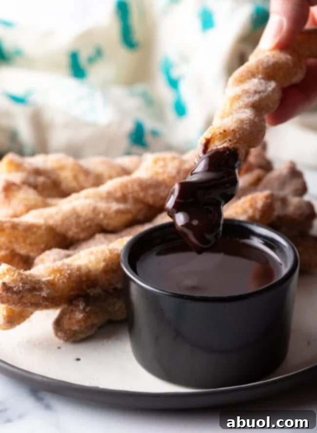 A hand dipping a crunchy, sugar-coated cinnamon twist into a small ramekin of rich chocolate sauce.