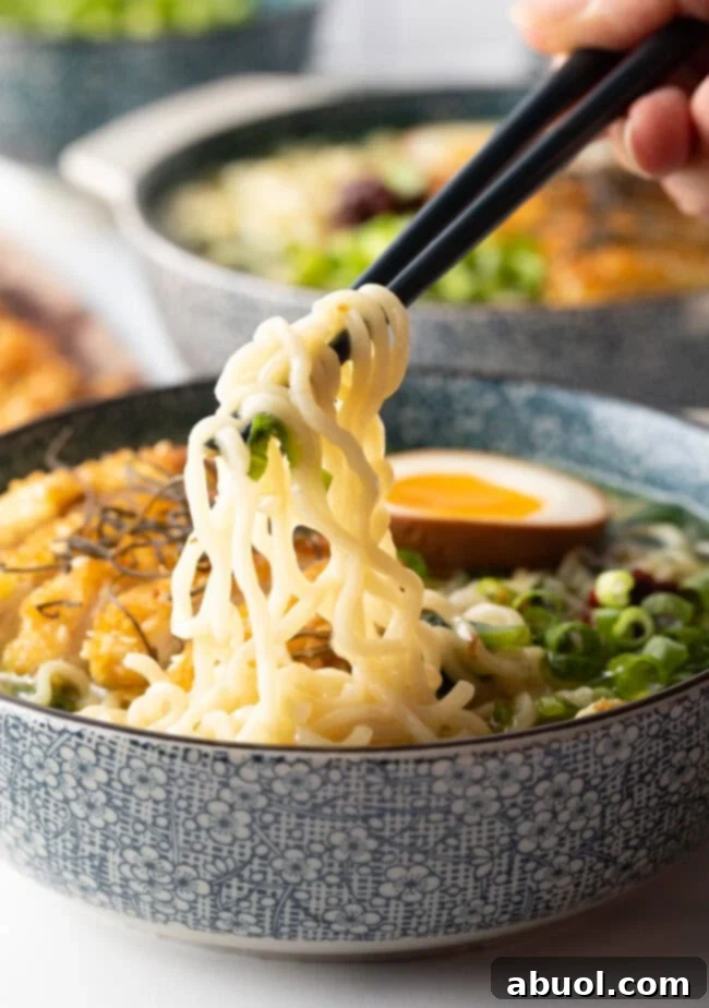 A hand holding chopsticks, lifting tender noodles from a generously filled soup bowl of chicken katsu ramen, enticingly garnished.