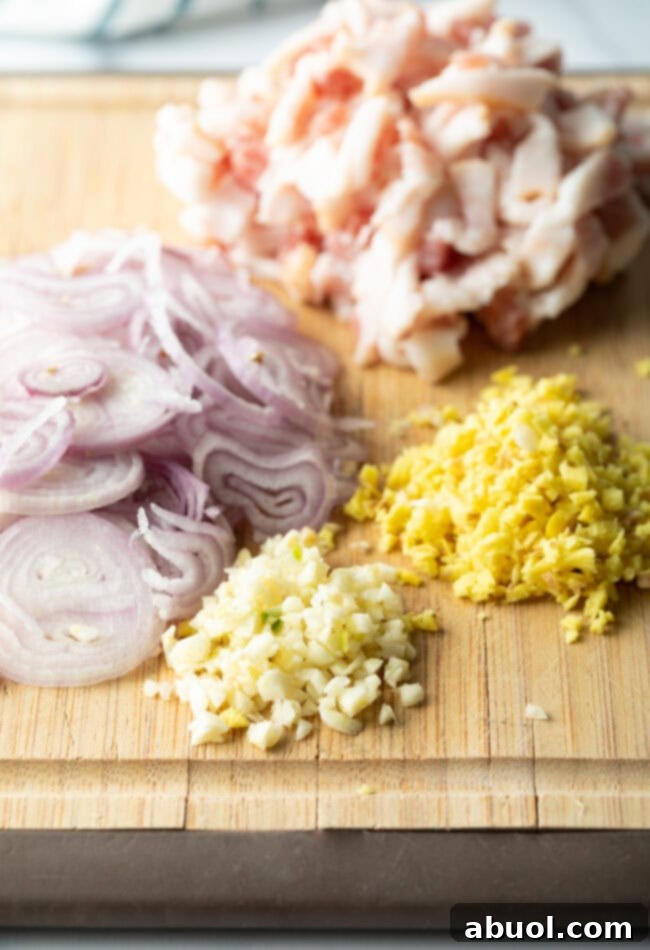 A wooden cutting board displaying fresh ingredients: minced garlic, grated ginger, thinly sliced shallots, and raw chicken cutlets.