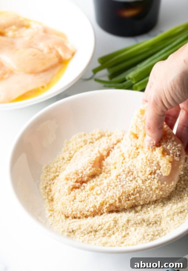 A hand carefully dipping a chicken cutlet into a bowl of seasoned breadcrumbs for coating.