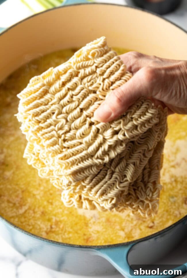 A hand holds blocks of instant ramen noodles, about to drop them into a pot of simmering, flavorful broth.