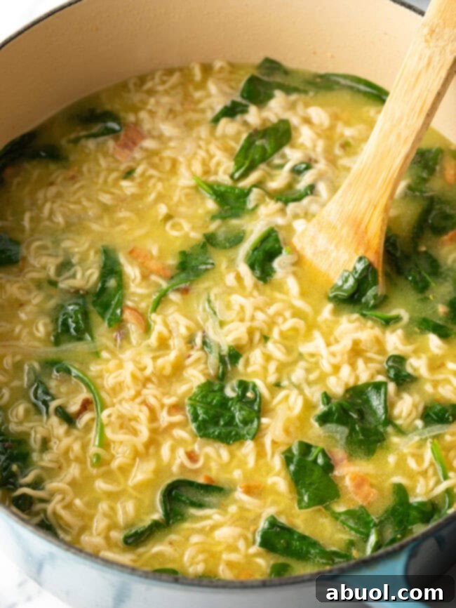 A wooden spoon stirring a rich katsu ramen broth in a pot, with noodles and greens visible.