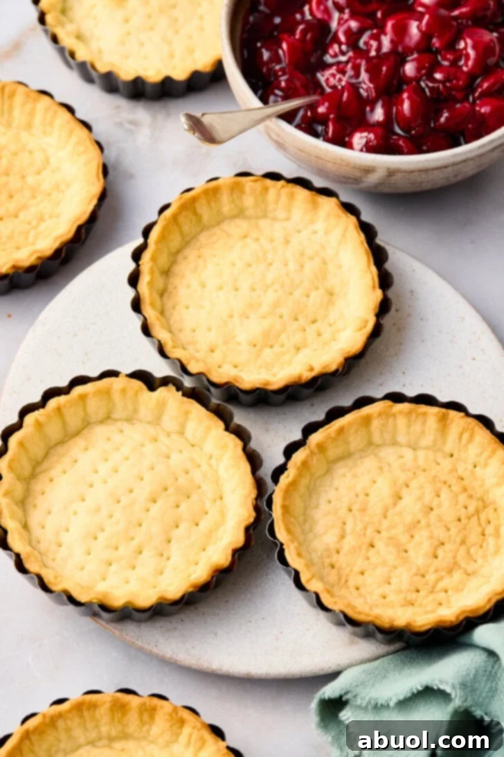 Baked cherry pie crusts in the tart pans next to a bowl of cherry pie filling. 
