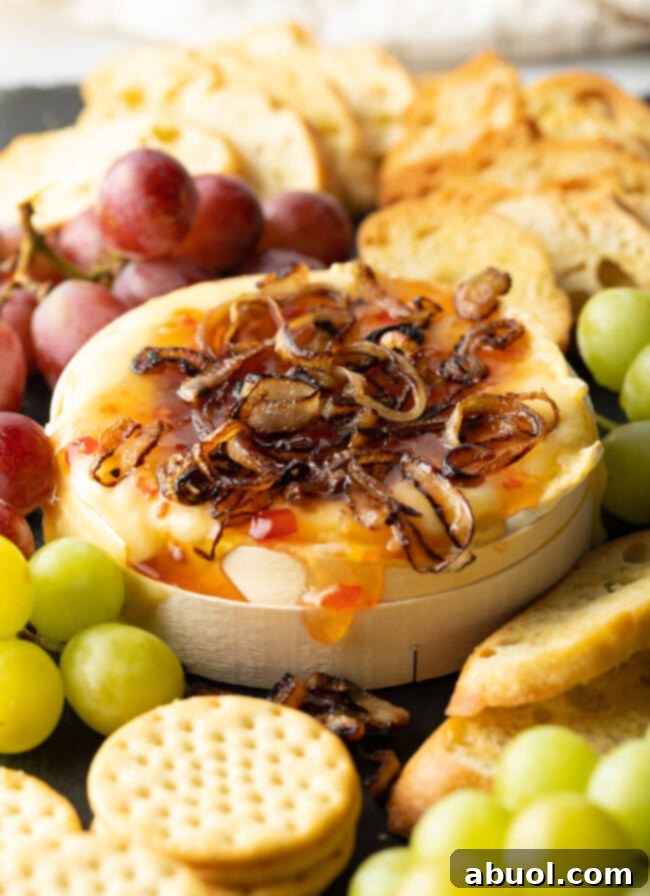Wheel of Camembert cheese baked with sweet chili sauce and sauteed shallots. It is on a platter and surrounded by red and green grapes, crackers, and baguette slices.