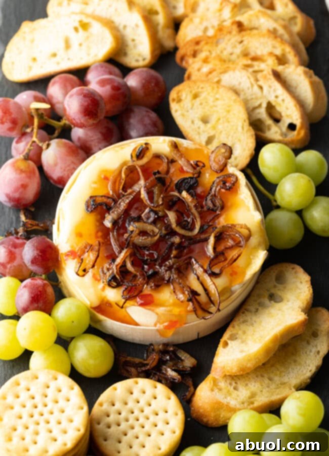 Wheel of Camembert cheese baked with sweet chili sauce and sauteed shallots. It is on a platter and surrounded by red and green grapes, crackers, and baguette slices.