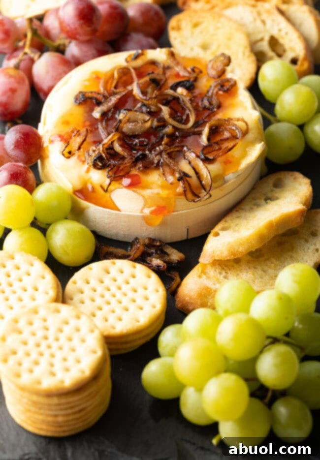 Wheel of Camembert cheese baked with sweet chili sauce and sauteed shallots. It is on a platter and surrounded by red and green grapes, crackers, and baguette slices.