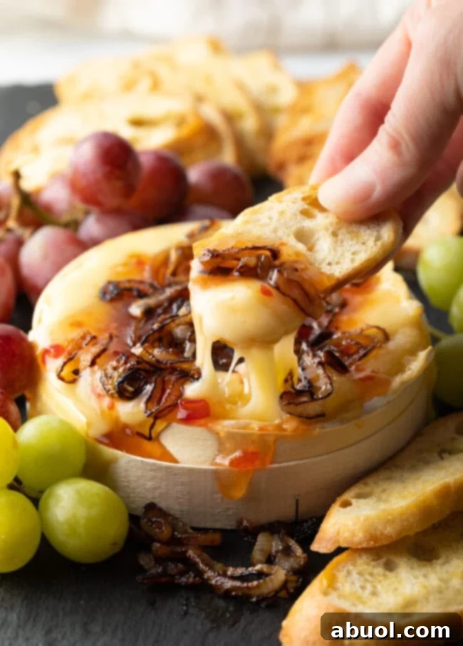 Hand scooping gooey melted cheese with onions and chili sauce from the baked wheel.