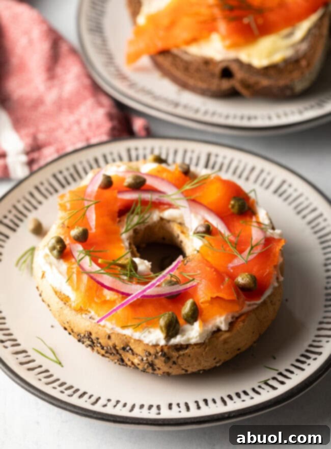 A perfectly prepared half bagel on a ceramic plate, generously topped with a creamy layer of cream cheese, artfully arranged slices of homemade lox (gravlax), scattered capers, and delicate rings of thinly sliced red onions.
