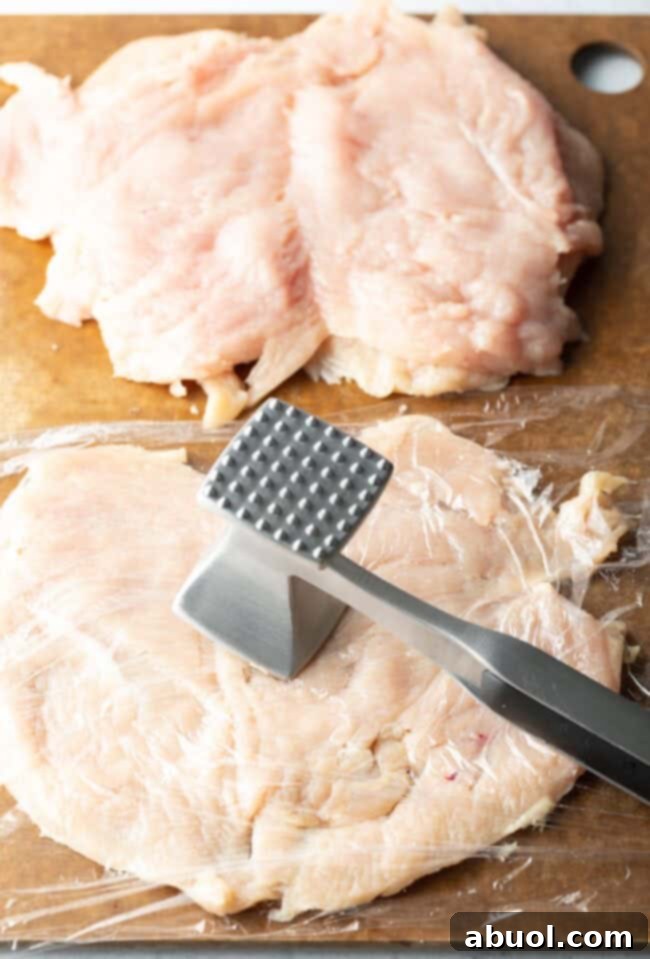 Butterflied chicken breasts on a cutting board, covered in plastic and with a metal mallet pounding them thin.