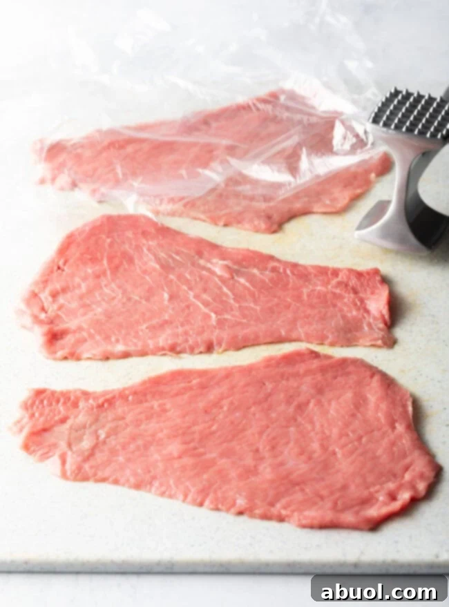 Four raw cutlets with plastic wrap and meat mallet. 