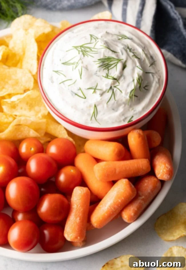 Top view bowl of dip with fresh chopped dill on top. Bowl is nestled on a plate with baby carrots, cherry tomatoes, and potato chips.