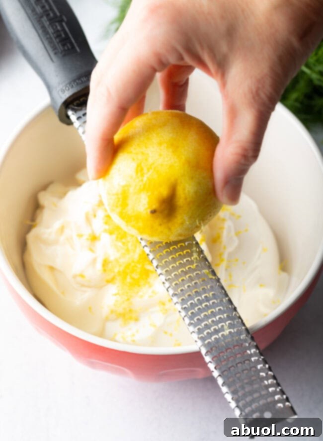Hand zesting lemon over bowl of sour cream.