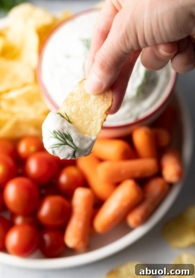 Hand with potato chip and dill dip, shown to camera hovering above plate with cherry tomatoes, baby carrots, and potato chips.