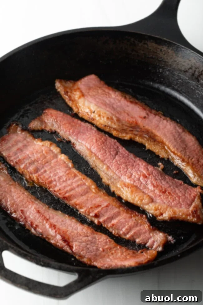 Four strips of beef bacon cooking in a cast iron skillet.