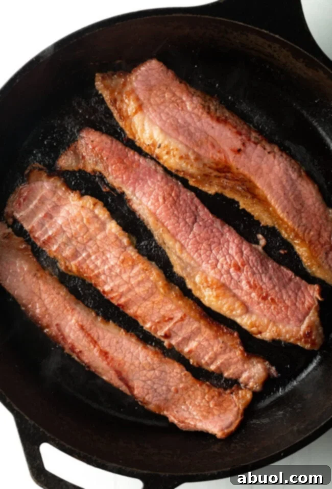 Top down view 4 pieces of bacon made with beef cooking in a cast iron skillet.