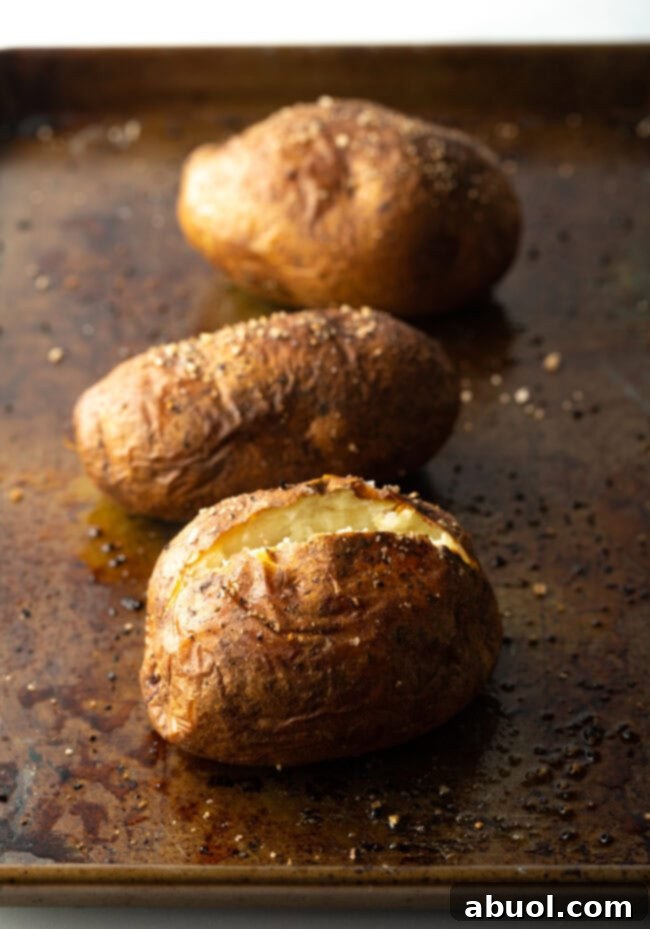 Three baked potatoes on a baking sheet. One closest to camera is split open at the top.