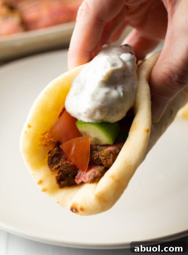 Savory Spiced Beef Shawarma 14 Close view hand holding flatbread naan beef shawarma recipe topped with chopped cucumbers, tomatoes, red onion and tzatziki sauce.