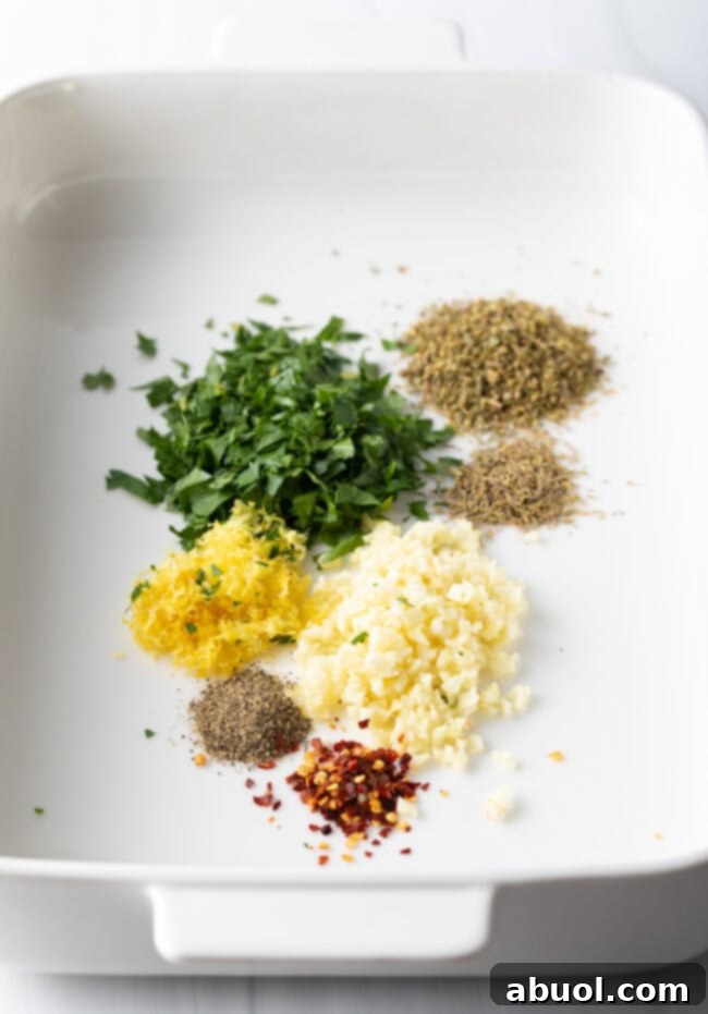 Various Greek spices, including oregano and thyme, laid out in a white baking dish, ready for mixing.