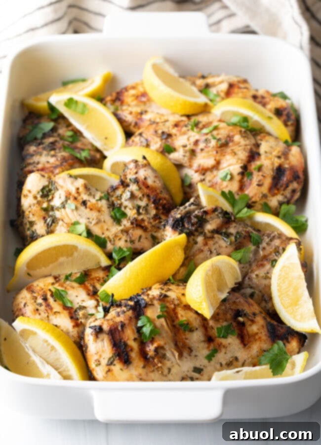 Side view of zestily marinated, grilled Greek chicken pieces with fresh lemon wedges, presented in a white baking dish.