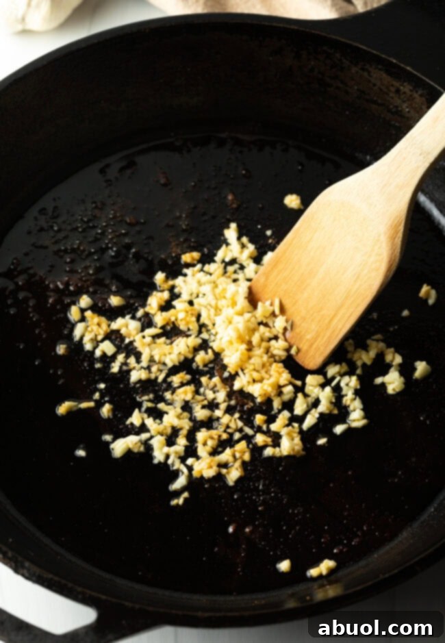 Minced garlic sizzling gently in a cast iron skillet, being stirred with a wooden spatula to prevent burning, creating a fragrant base for the glaze.