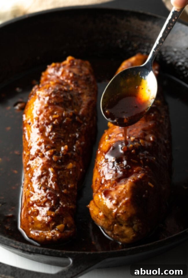 A hand spooning additional honey garlic glaze over two baked pork tenderloins in a cast iron skillet, enhancing the shine and flavor.