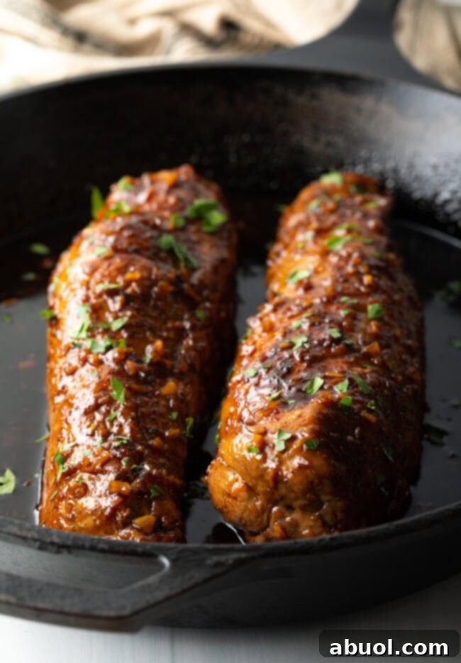 Two glistening oven-roasted pork tenderloins, beautifully glazed and resting in a cast iron skillet after baking, ready to be sliced.