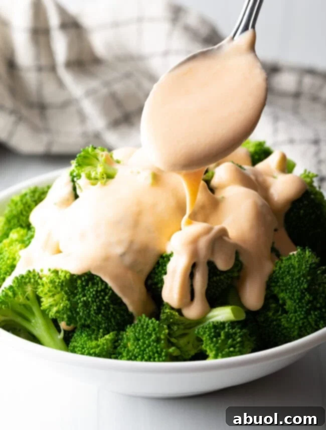 Bowl of steamed broccoli with spoon pouring rich, smooth cheese sauce on top.