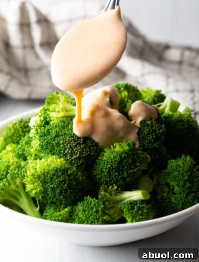 Metal spoon gracefully pouring smooth, golden cheese sauce over freshly steamed broccoli florets.