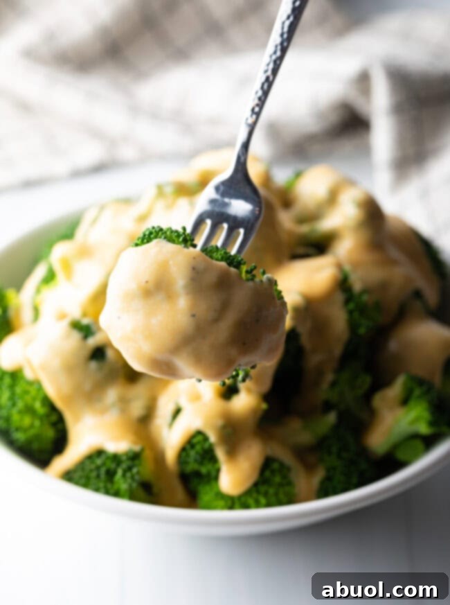 Fork holding a perfect floret of broccoli, richly coated with smooth, homemade cheese sauce, presented to the camera.