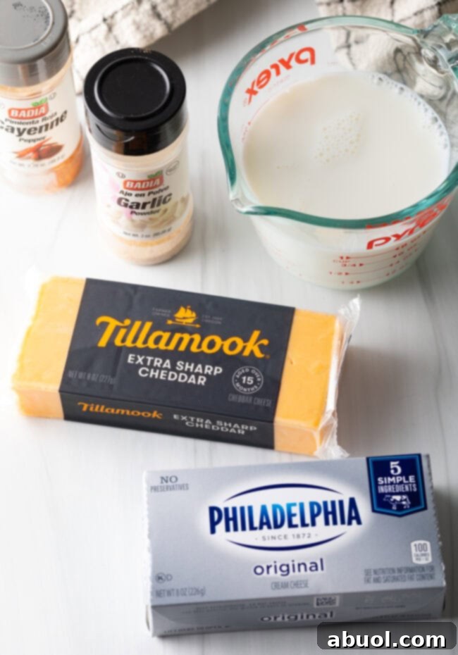 Recipe ingredients laid out on a white cutting board: a block of sharp cheddar cheese, a block of cream cheese, a glass measuring cup with milk, and small bottles of garlic and cayenne powder spices.