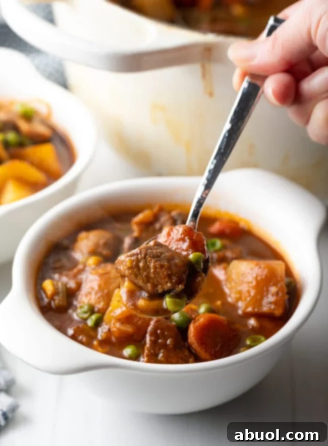 Mulligan beef stew with chopped potatoes, carrots, peas, and chunks of meat in a white bowl. Hand with metal spoon showing a spoonful to camera.