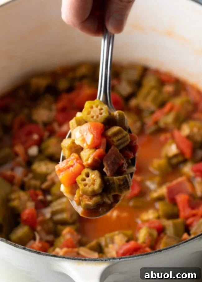 A hand holding a metal spoon, showcasing a perfect spoonful of tomatoes and okra stew towards the camera, highlighting its rustic appeal.