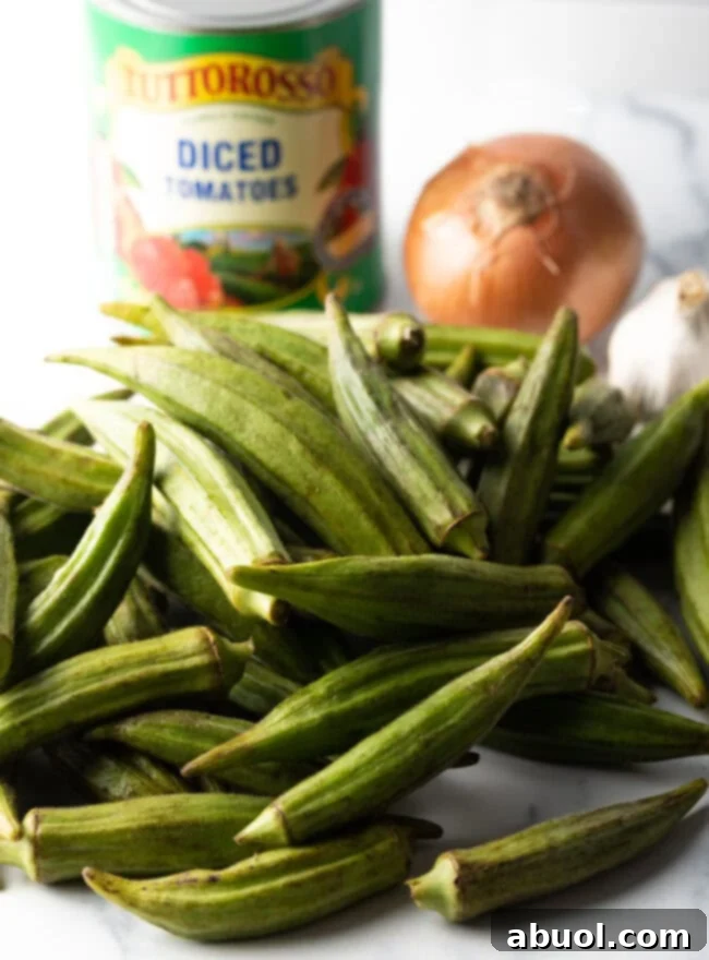 Close-up shot of fresh ingredients: a pile of vibrant green okra pods on a cutting board, a can of diced tomatoes, and a whole yellow onion, ready for preparation.