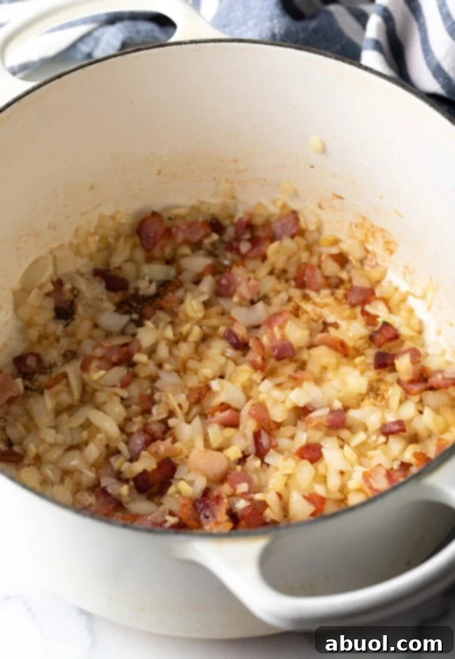 Diced onion and crispy bacon cooking in a pristine white Dutch oven, showcasing the initial stages of building flavor.
