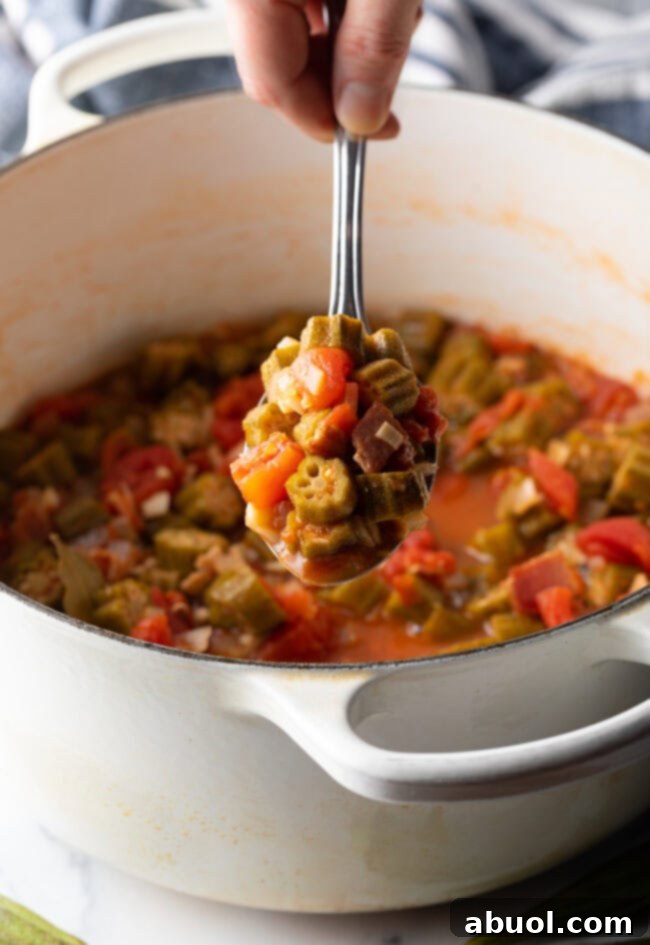 A hand holding a metal spoon, presenting a generous spoonful of the finished tomatoes and okra stew, rich with flavor.