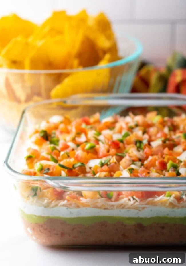 Glass square baking dish with layered Mexican bean dip. A bowl of tortilla chips is in the background.
