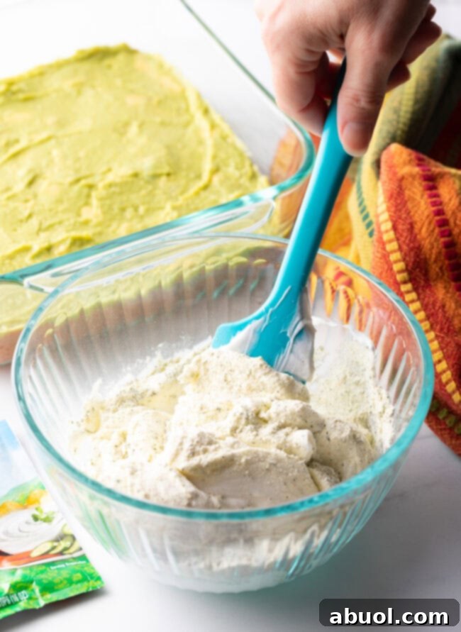 Hand with blue spatula, blending sour cream layer ingredients together in a small glass bowl.