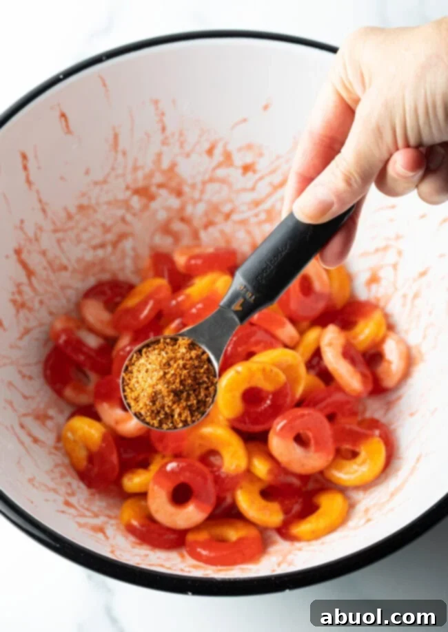 Hand adding Tajin seasoning from a metal measuring spoon into a bowl of gummy rings.