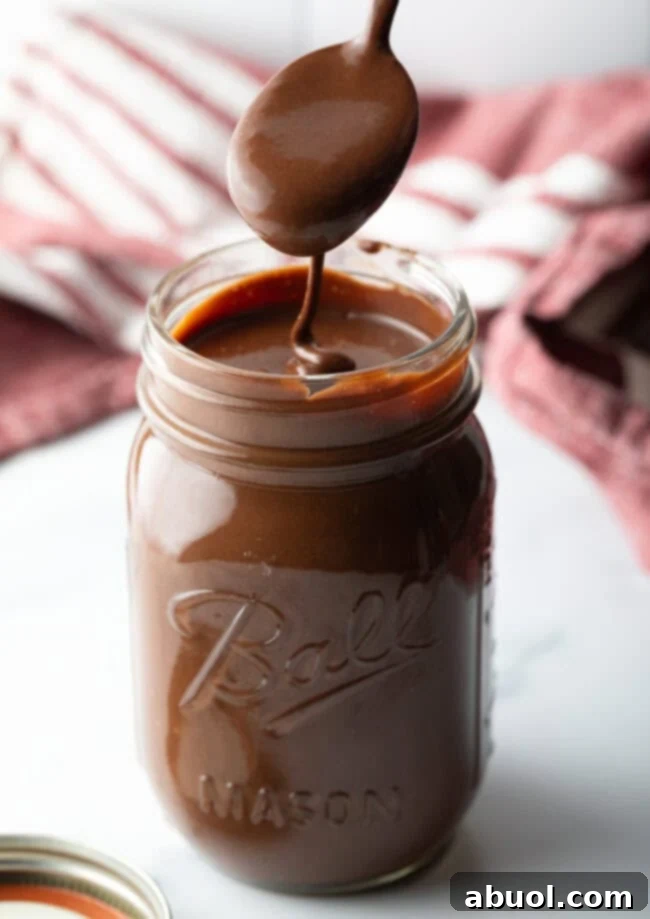 Metal spoon pouring the hot fudge sauce into a glass mason jar.